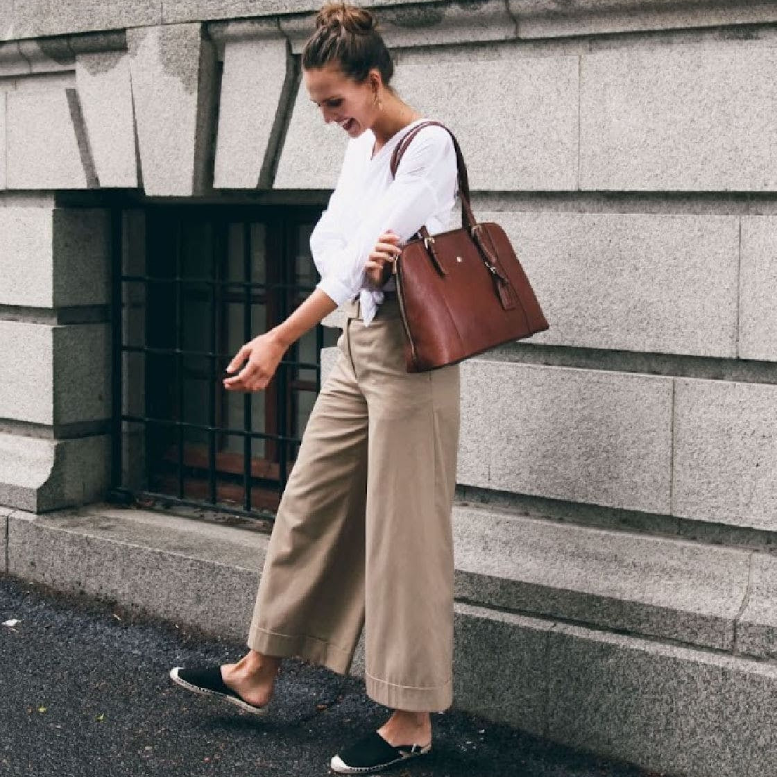 Woman holding a brown leather tote bag against a stone wall.