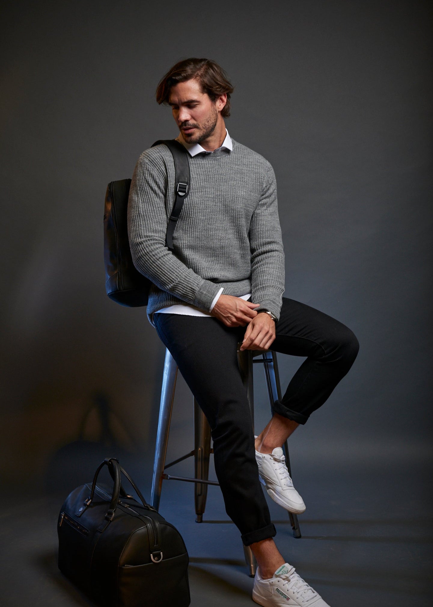 Man sitting on a stool with a backpack and suitcase against a dark background