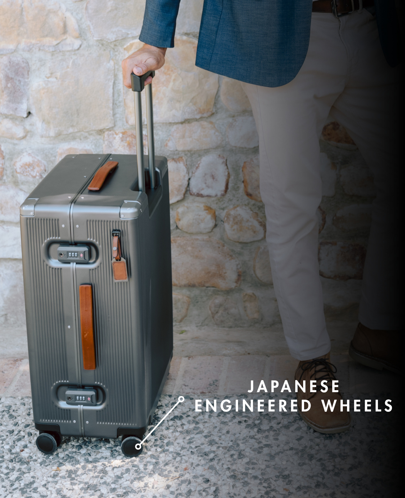 Person holding a gray suitcase with 'Japanese Engineered Wheels' text against a stone wall.