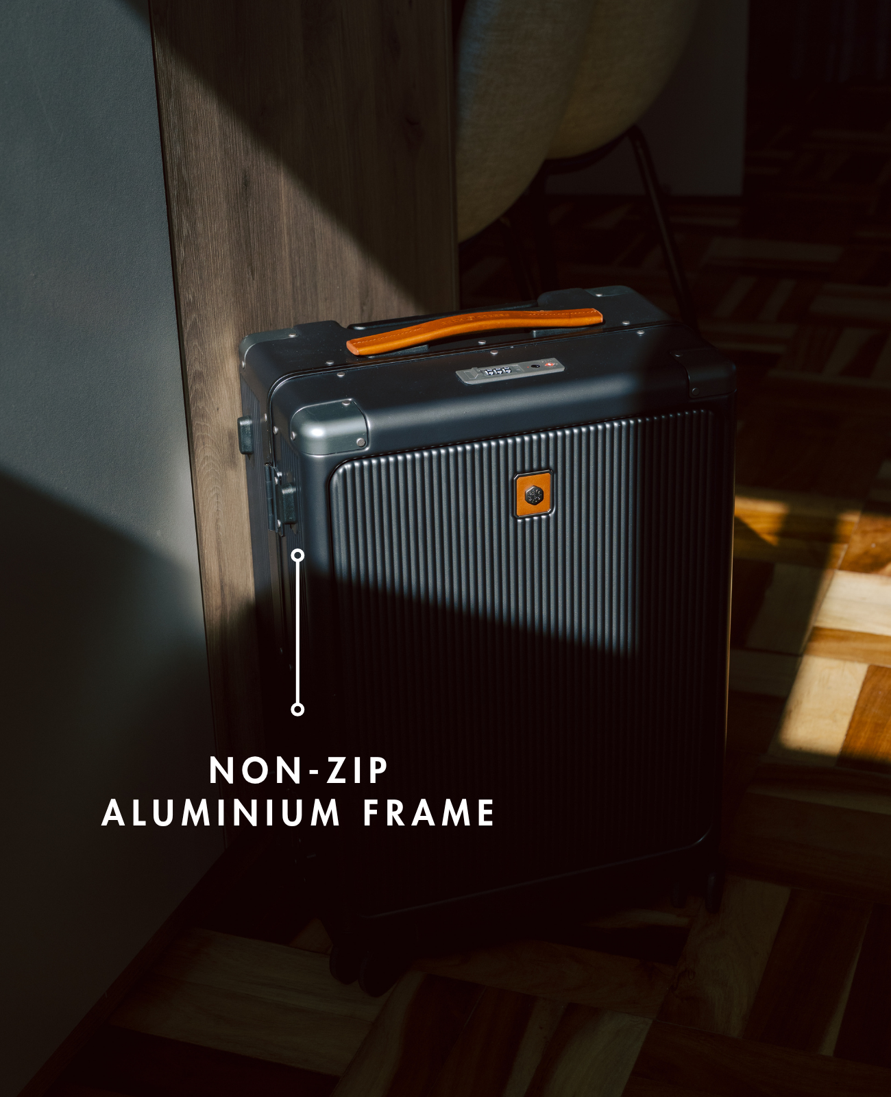 Black suitcase with orange handle on a wooden floor, labeled 'Non-Zip Aluminium Frame'.