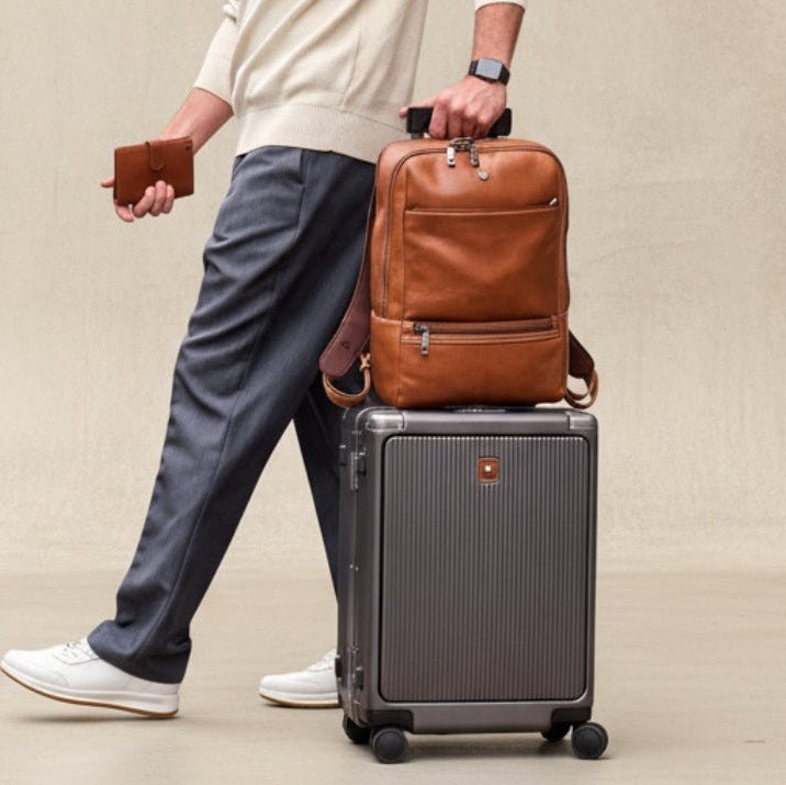 Person holding a brown leather backpack and a gray suitcase on a beige background
