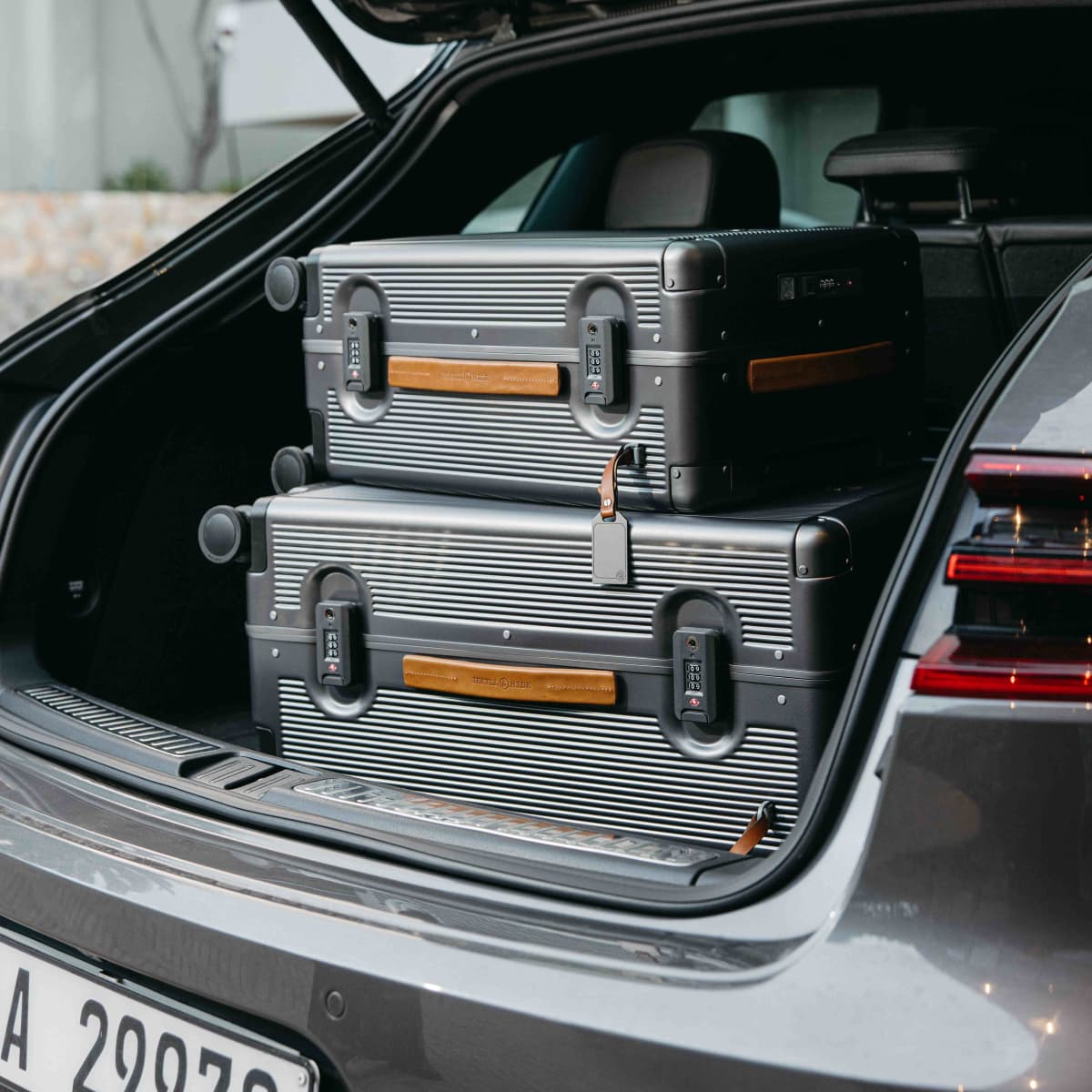 Two hard-shell silver suitcases stacked in the open trunk of a gray car, with a leatherette handle and small luggage tag visible.