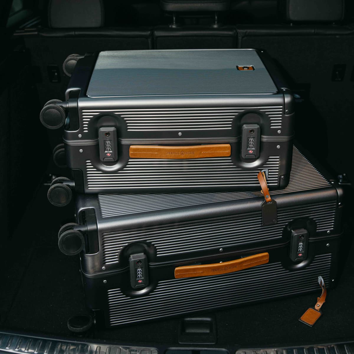 Two hard-shell spinner suitcases stacked inside a dark car trunk. The suitcases are dark gray with horizontal ribbing, black combination locks, and tan leather handles and luggage tags.