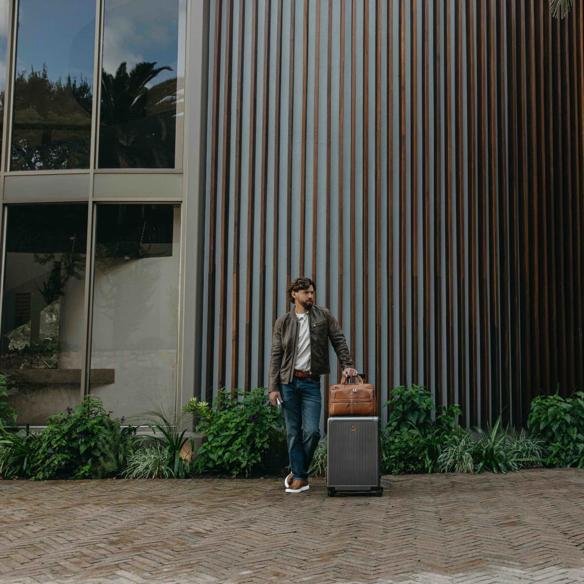 A man wearing a brown leather jacket, white shirt, and blue jeans stands outdoors next to a dark gray rolling suitcase, holding a brown leather duffel bag. He is positioned in front of a modern building with vertical brown metal siding and green plants along the base.