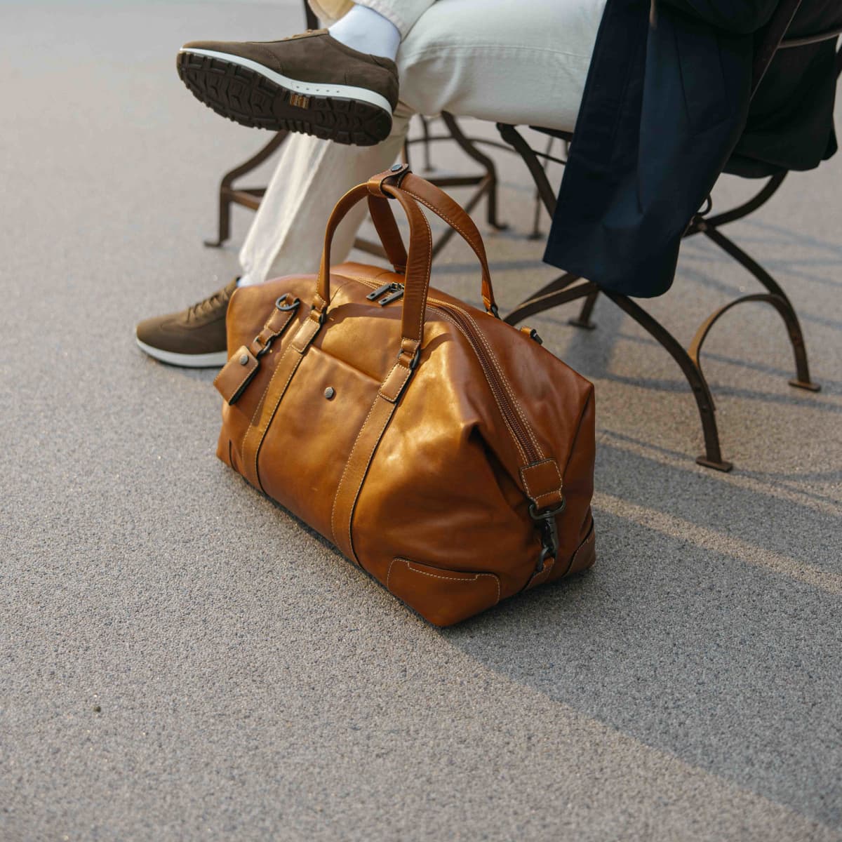 A tan leather duffel bag with dual top handles and a zippered closure, placed on a gray paved surface next to a person sitting on a metal chair.