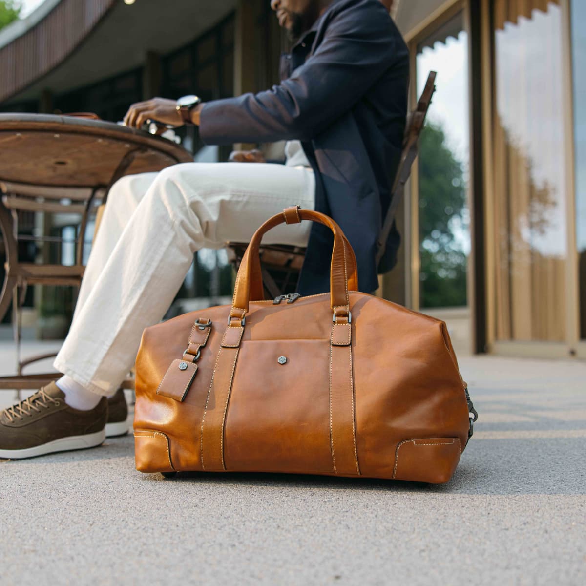 A tan leather duffel bag sits on a concrete surface in front of a seated man at an outdoor cafe table. The man is wearing a navy blazer, off-white trousers, and brown shoes.
